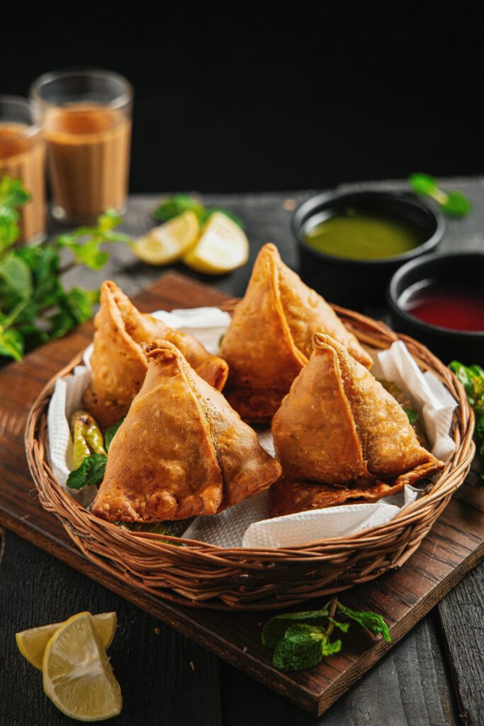 Fried golden samosas on white tissues in wicker basket on board surrounded by lemon and salad dressing in deep dishes and beverages in glass