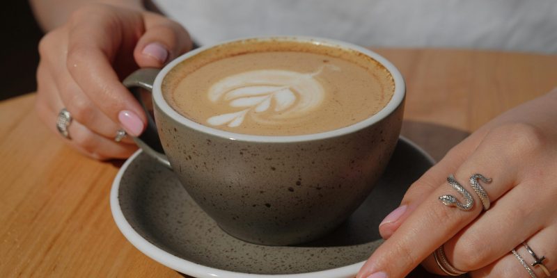 A close-up shot of hands with rings holding a cappuccino featuring delicate latte art on a wooden table.